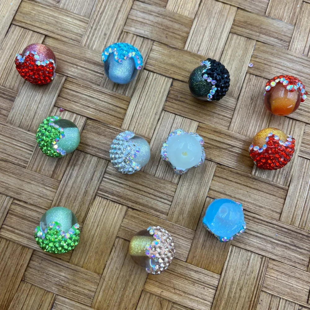 Assorted rhinestone-encrusted galaxy glitter resin fancy beads in red, blue, green, black, orange, white, and gold colorways displayed on woven bamboo surface for DIY jewelry making and beadable pens.
