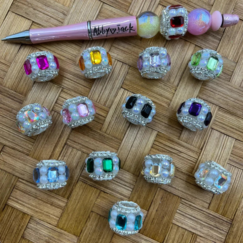 Assorted rhinestone-bordered fancy beads with large center gemstones and opalescent pearl accent beads in pink, yellow, purple, green, black, blue, teal, and emerald colors, displayed on a woven surface next to a pink beadable pen labeled Abby and Jack.