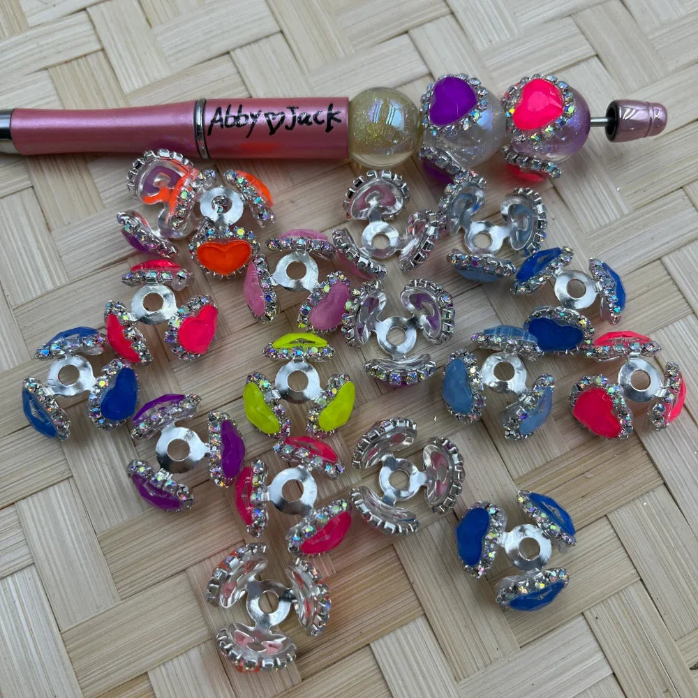 Assorted colorful rhinestone heart bead caps with silver crystal trim in neon pink, blue, orange, purple, and yellow, displayed next to a beadable pen on a wicker surface.