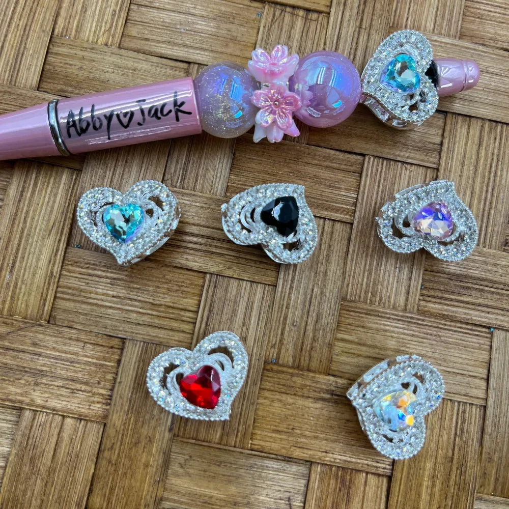 Six decorative silver rhinestone heart-shaped snap charms with colorful center gemstones in blue, black, purple, red, and iridescent AB crystal, displayed on a woven rattan surface next to a pink beadable pen decorated with matching heart charm.
