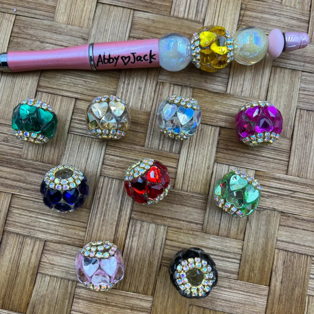 Assorted rhinestone-rimmed heart faceted crystal fancy beads in green, gold, iridescent, magenta, navy, red, light green, pink, and black displayed on a woven surface next to a decorative beadable pen labeled Abby Jack.