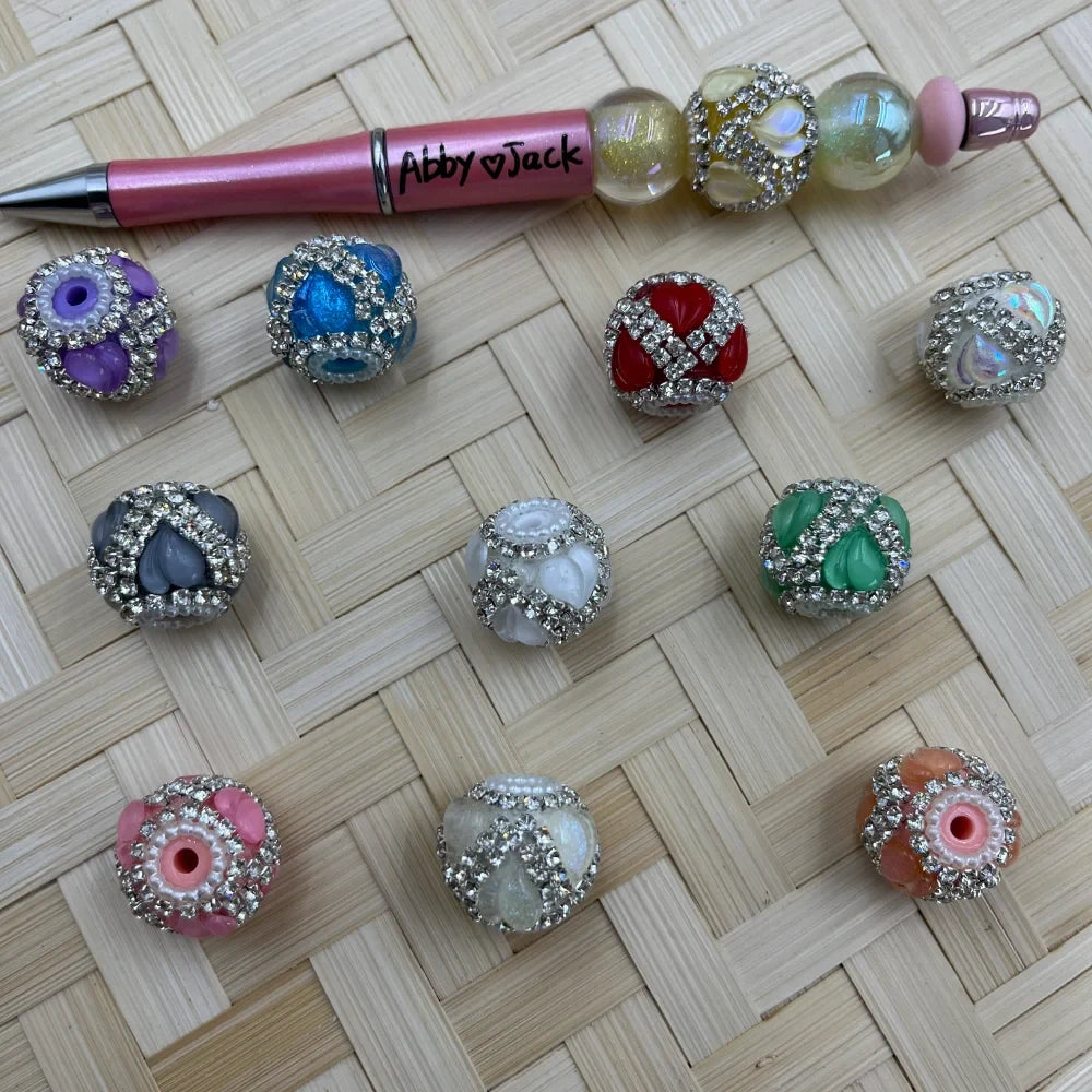 Assorted rhinestone-encrusted heart fancy beads in purple, blue, red, iridescent, gray, white, green, pink, and peach colors displayed on a woven surface alongside a pink beadable pen labeled Abby Jack.