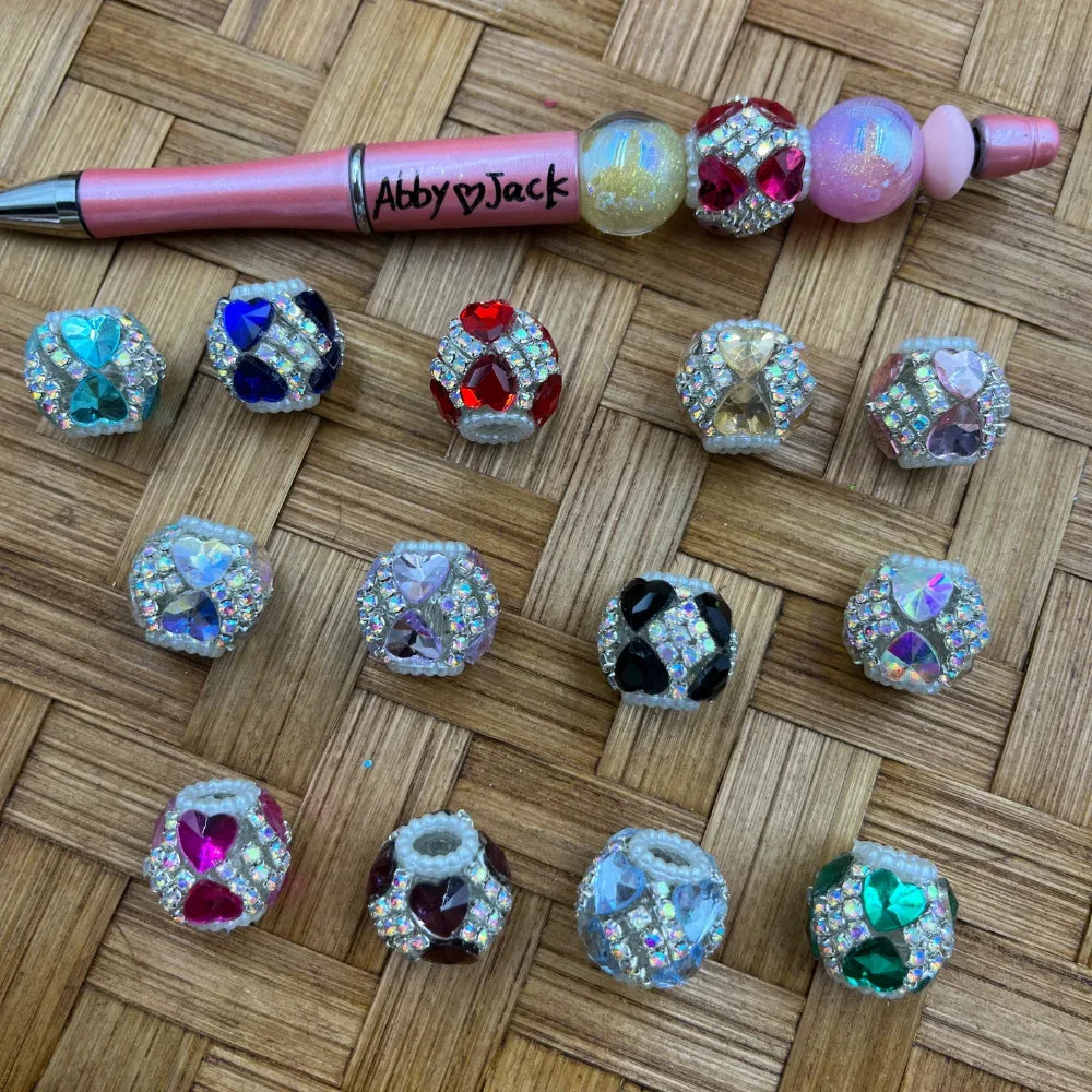Assorted rhinestone heart gem fancy beads in multiple colors including teal, blue, red, pink, purple, black, and green with crystal rhinestone accents, displayed alongside a beadable pen on a woven surface.