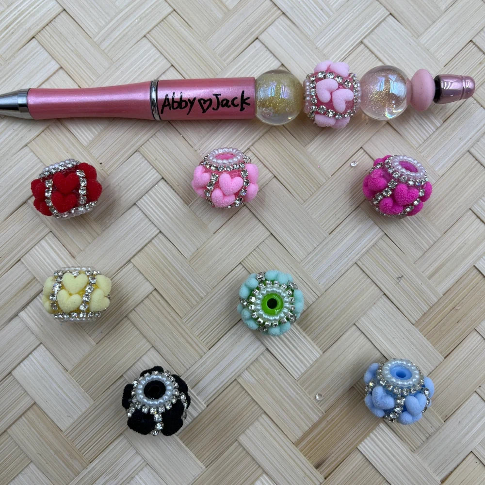 Assorted rhinestone-trimmed fancy beads featuring heart and pom pom textures in red, pink, magenta, yellow, teal, black, and blue colors displayed on a woven surface next to a pink beadable pen branded Abby Jack.