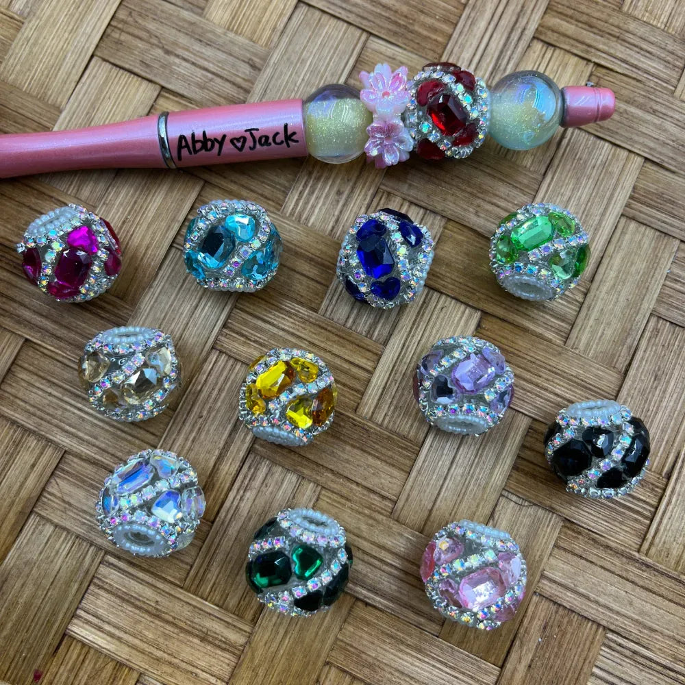 Assorted rhinestone-encrusted large gem fancy beads in multiple colors including pink, teal, blue, green, yellow, purple, black, and clear, displayed on woven surface next to a decorated beadable pen labeled Abby Jack.
