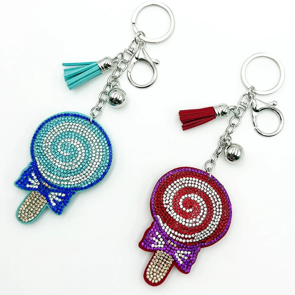 Two rhinestone-studded lollipop charm keychains in teal/blue and red/purple with tassels, silver rings, and bell accents for DIY jewelry making and keychains.