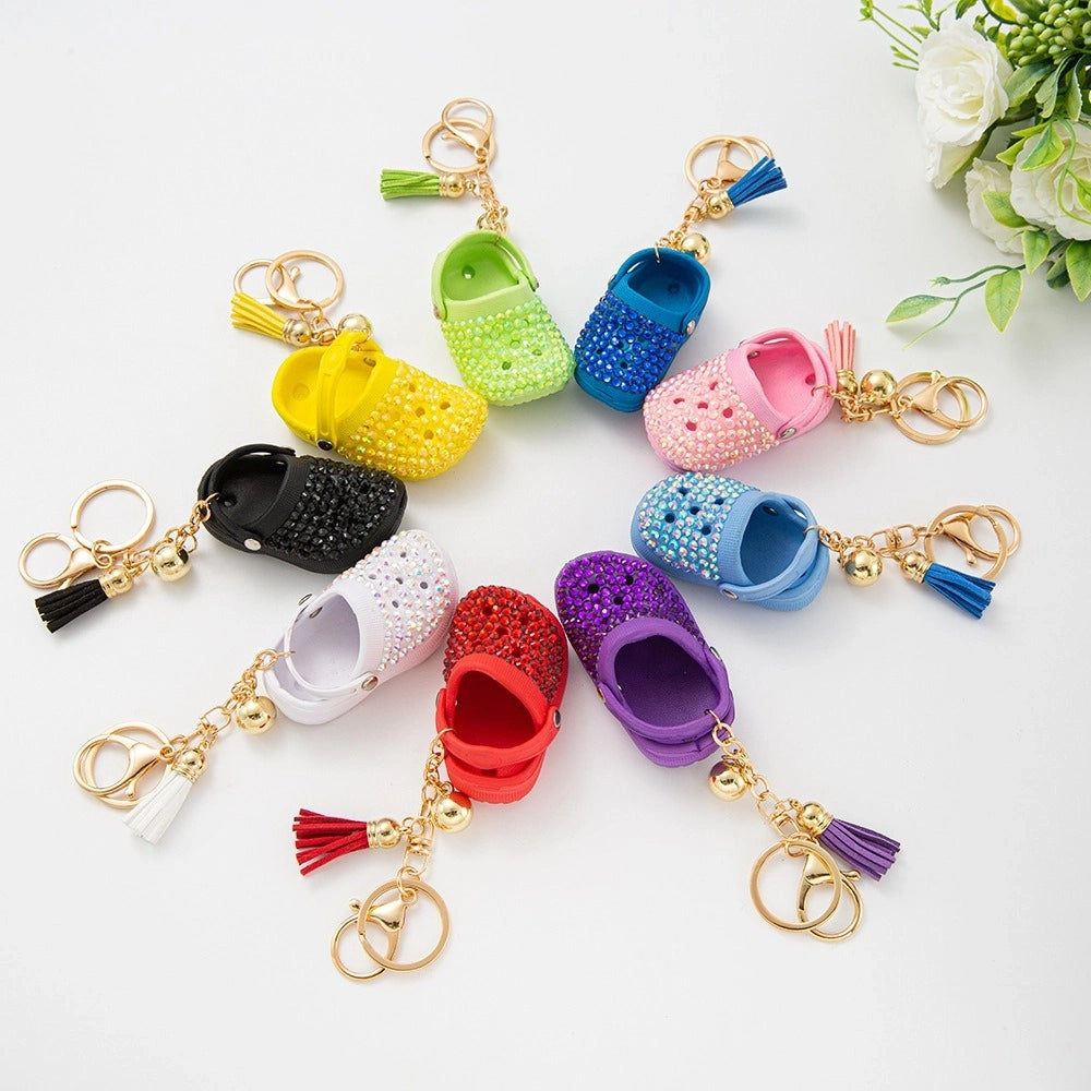 Eight colorful rhinestone-encrusted mini croc shoe keychain charms with gold chain and tassel in yellow, green, blue, pink, lavender, red, white, black, and purple arranged in a circle on a white surface.