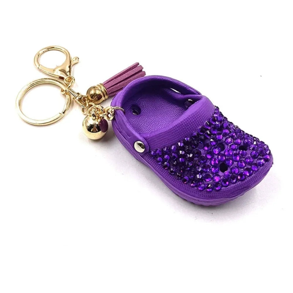 Eight colorful rhinestone-encrusted mini croc shoe keychain charms with gold chain and tassel in yellow, green, blue, pink, lavender, red, white, black, and purple arranged in a circle on a white surface. - Purple