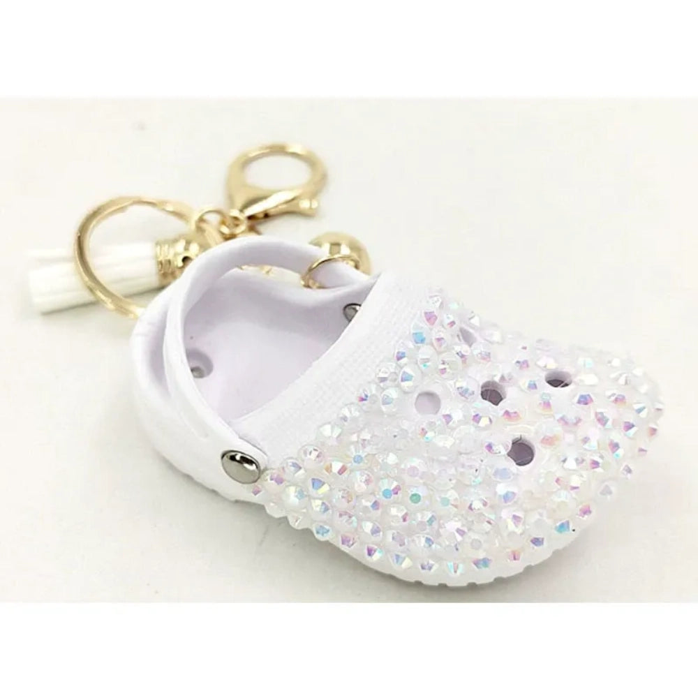 Eight colorful rhinestone-encrusted mini croc shoe keychain charms with gold chain and tassel in yellow, green, blue, pink, lavender, red, white, black, and purple arranged in a circle on a white surface. - White