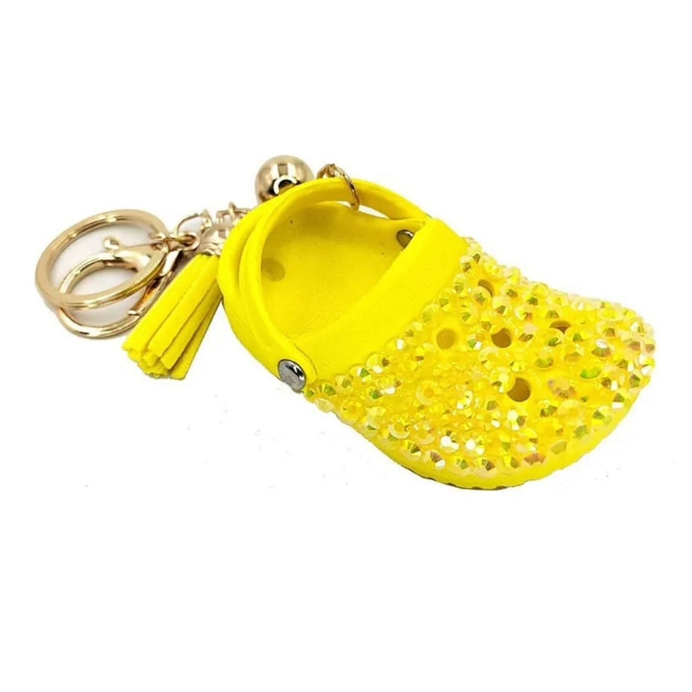 Eight colorful rhinestone-encrusted mini croc shoe keychain charms with gold chain and tassel in yellow, green, blue, pink, lavender, red, white, black, and purple arranged in a circle on a white surface. - Yellow