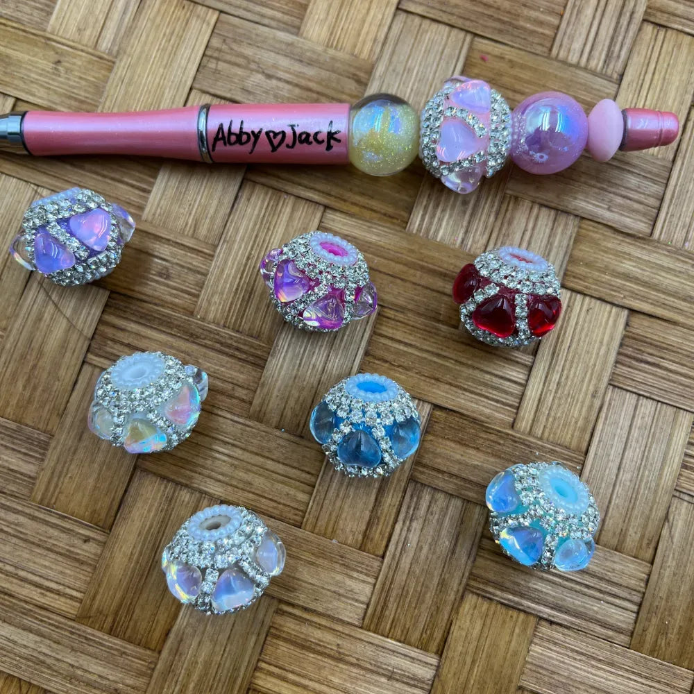 Assorted rhinestone-encrusted fancy beads with iridescent and colored heart crystal accents in purple, pink, red, blue, and opal white, displayed alongside a decorated beadable pen on a wicker surface.