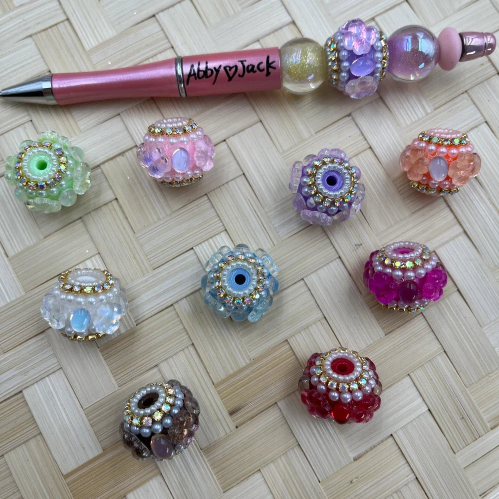 Assorted rhinestone and pearl cluster fancy beads in pastel colors including pink, purple, green, blue, red, peach, white, and brown displayed on a wicker surface next to a decorated beadable pen labeled Abby Jack.