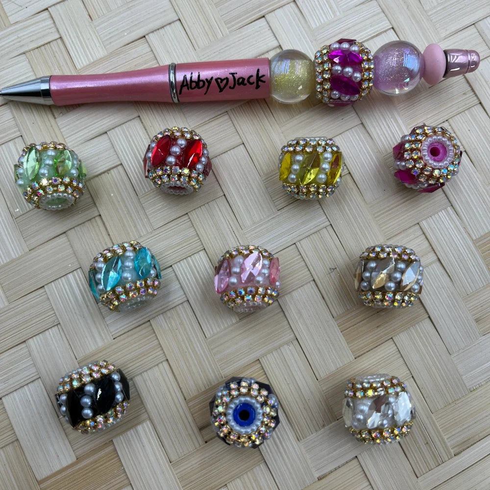 Assorted rhinestone and pearl embellished crystal fancy beads in multiple colors including red, yellow, pink, teal, black, clear, and green, displayed next to a decorative beaded pen on a woven surface.