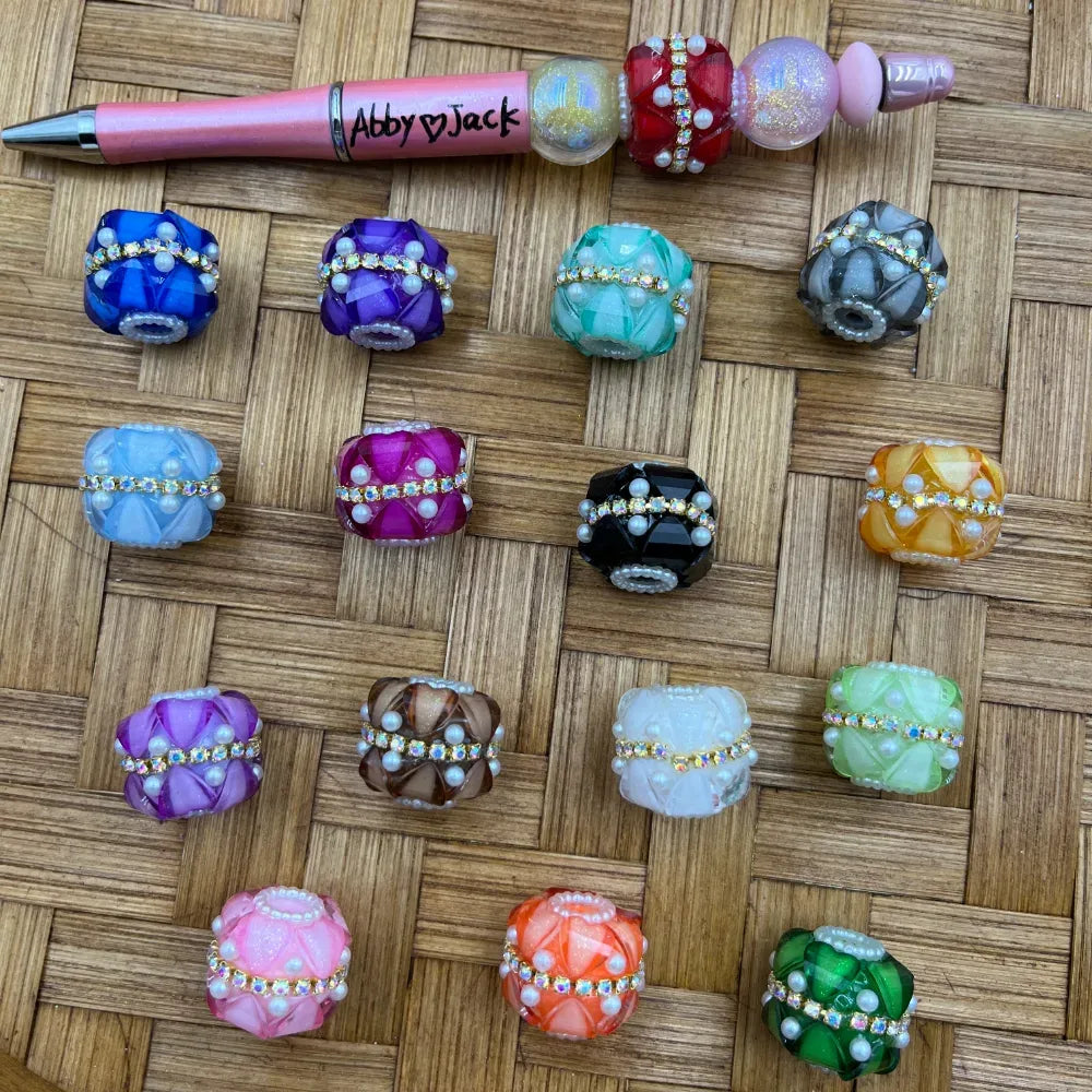 Assorted rhinestone and pearl-trimmed fancy chunky beads in multiple colors including blue, purple, teal, gray, black, pink, orange, and green displayed on a wicker surface next to a beadable pen.