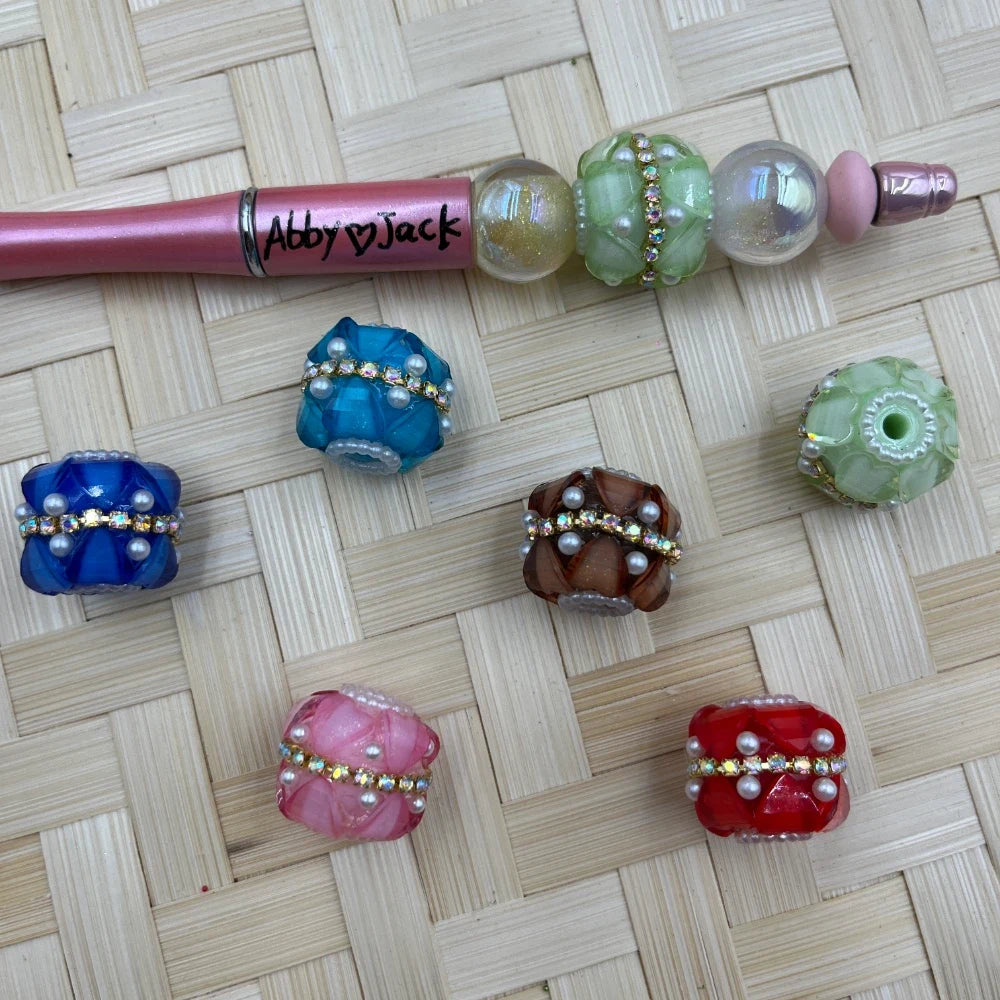 Assorted colorful chunky fancy beads with rhinestone crystal band and pearl accents in blue, teal, green, pink, red, and brown, displayed with a pink beadable pen on woven bamboo surface.