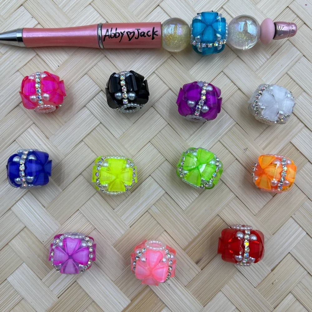 Assorted rhinestone and pearl flower cube fancy beads in 12 colors including pink, black, purple, white, blue, yellow-green, green, orange, magenta, light pink, and red, displayed on a woven bamboo surface next to an Abby Jack beadable pen.
