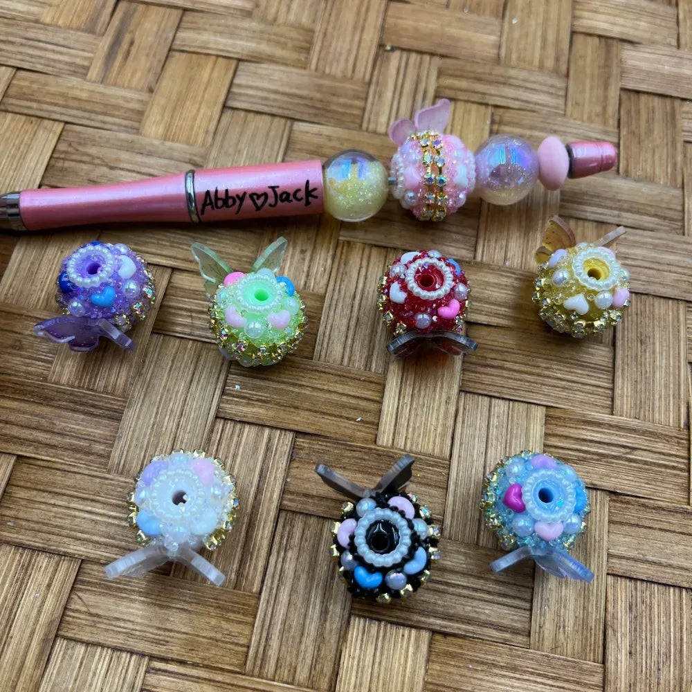 Assorted rhinestone and pearl flower fancy beads in purple, pink, red, yellow, black, and white with crystal accents displayed on a woven surface next to a pink beadable pen labeled Abby Jack.