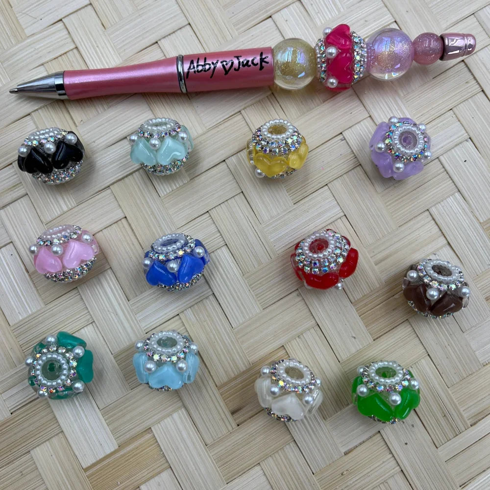 Assorted rhinestone and pearl-encrusted flower chunky fancy beads in 12 colors including black, mint, yellow, lavender, pink, blue, red, brown, teal, white, and green, displayed on a woven surface next to a pink beadable pen branded Abby and Jack.