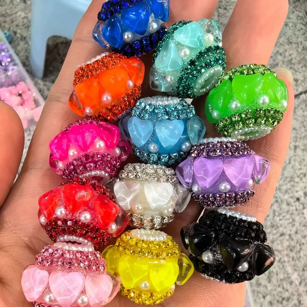 Assorted colorful rhinestone and pearl embellished heart flower acrylic fancy beads in multiple colors including blue, teal, green, orange, pink, red, purple, white, yellow, and black held in a hand.
