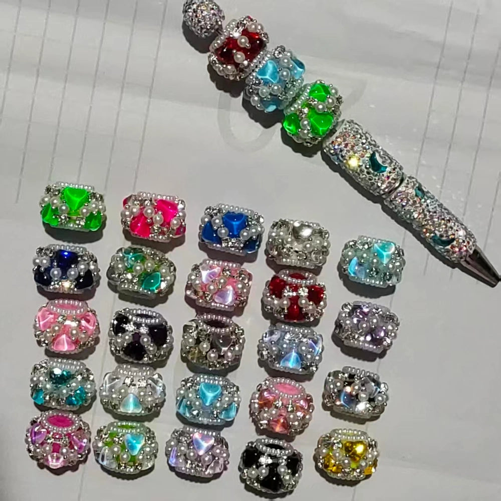 Assorted colorful square fancy beads decorated with rhinestones, faux pearls, and heart-shaped gemstones in multiple colors including red, green, blue, pink, black, and yellow, displayed next to a bejeweled beadable pen on white lined paper.