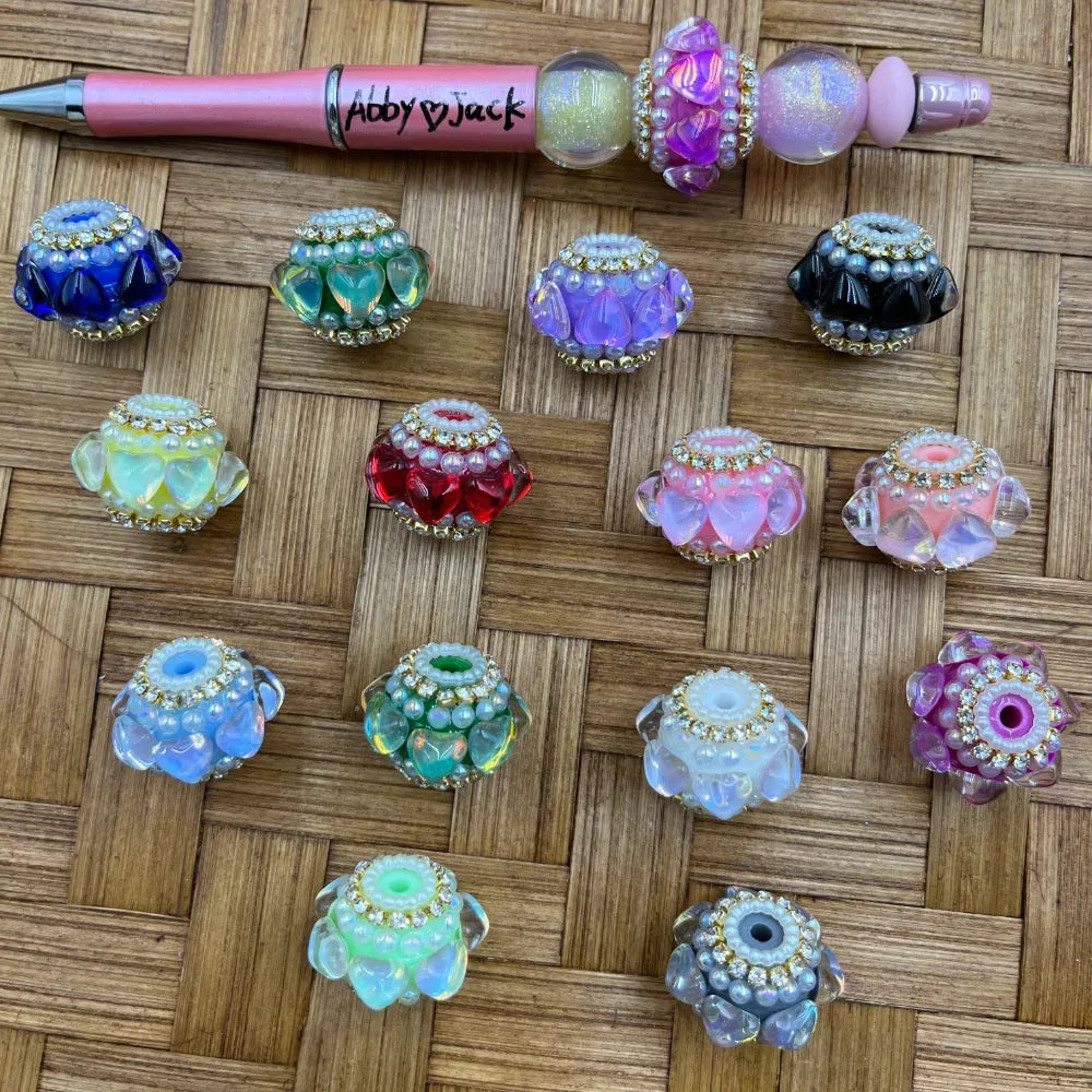 Assorted rhinestone and pearl heart petal fancy beads in 14 colors including blue, green, red, purple, pink, black, white, and clear displayed on a wicker surface next to a pink beadable pen branded Abby Jack.