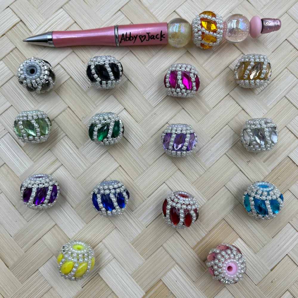 Assorted rhinestone and pearl-trimmed marquise gem fancy beads in multiple colors including pink, green, purple, blue, red, teal, and yellow, displayed on a woven surface next to a beadable pen.