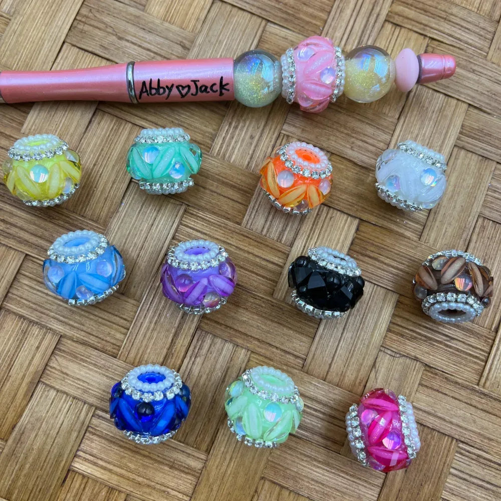 Assorted rhinestone and pearl rimmed faceted fancy beads in 12 colors including blue, purple, orange, black, pink, green, yellow, and white, displayed on a wicker surface next to a decorated beadable pen branded Abby Jack.