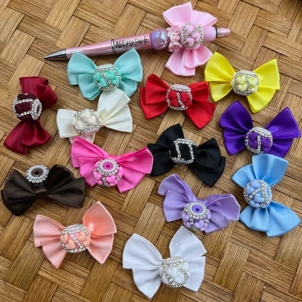 Collection of colorful satin ribbon bows with rhinestone and pearl fancy beads in assorted colors including pink, red, black, purple, blue, white, and more, displayed on a woven bamboo surface alongside a decorative beadable pen.