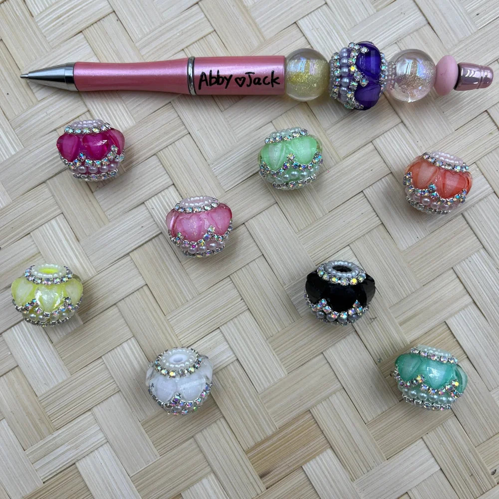 Assorted rhinestone and pearl-trimmed fancy chunky beads in pink, green, black, white, coral, mint, and yellow colors displayed on a woven surface beside a decorative beadable pen labeled Abby and Jack.