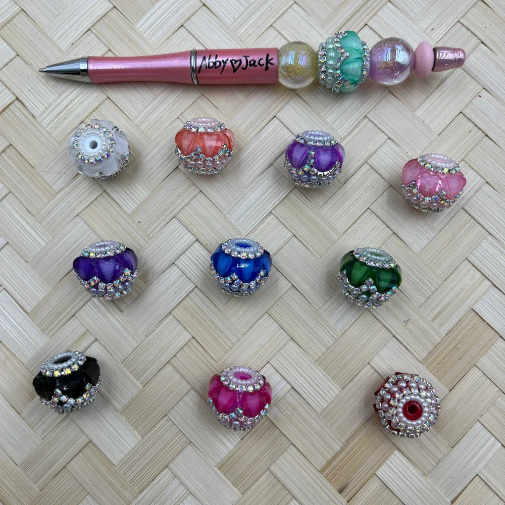 Assorted rhinestone and pearl-trimmed fancy chunky beads in multiple colors including black, pink, purple, blue, green, coral, and hot pink, displayed on a woven surface next to a decorated beadable pen.