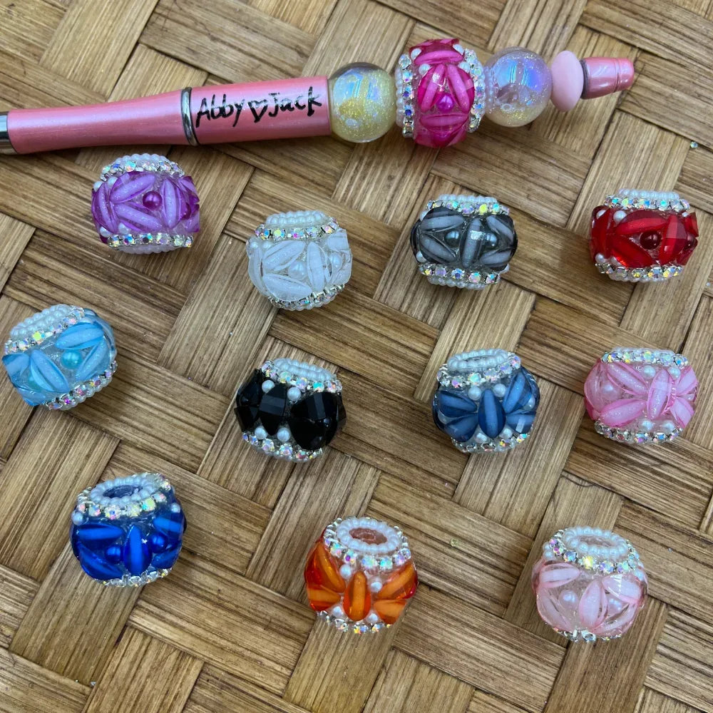 Assorted rhinestone-trimmed petal fancy beads in multiple colors including purple, white, gray, red, blue, black, pink, orange, and royal blue, displayed on a wicker surface alongside a decorated beadable pen labeled Abby and Jack.