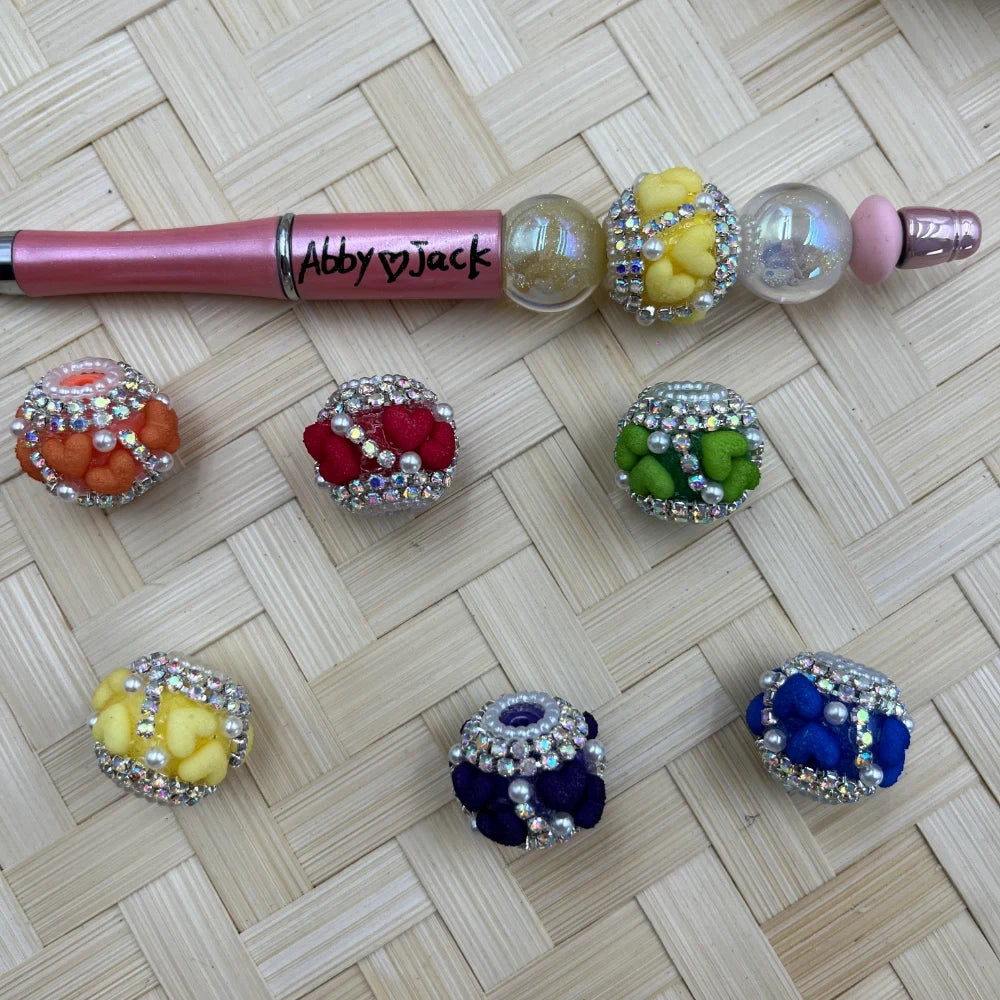 Assorted rhinestone-trimmed pom pom fancy beads in orange, red, green, yellow, purple, and blue with pearl accents, shown with a pink beadable pen on woven background.