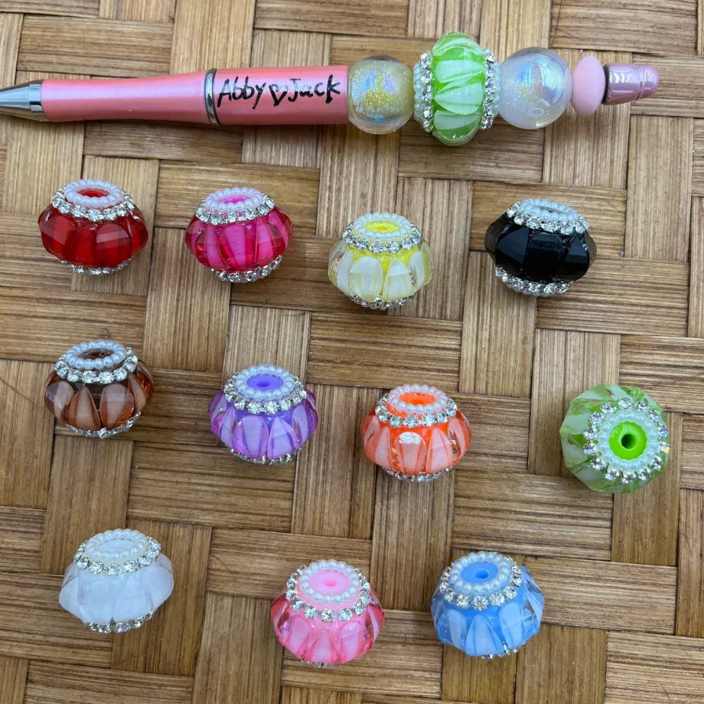 Assorted rhinestone-rimmed faceted resin fancy beads in multiple colors including red, pink, yellow, black, orange, purple, blue, green, and white, displayed on a woven surface next to a decorated beadable pen labeled Abby and Jack.