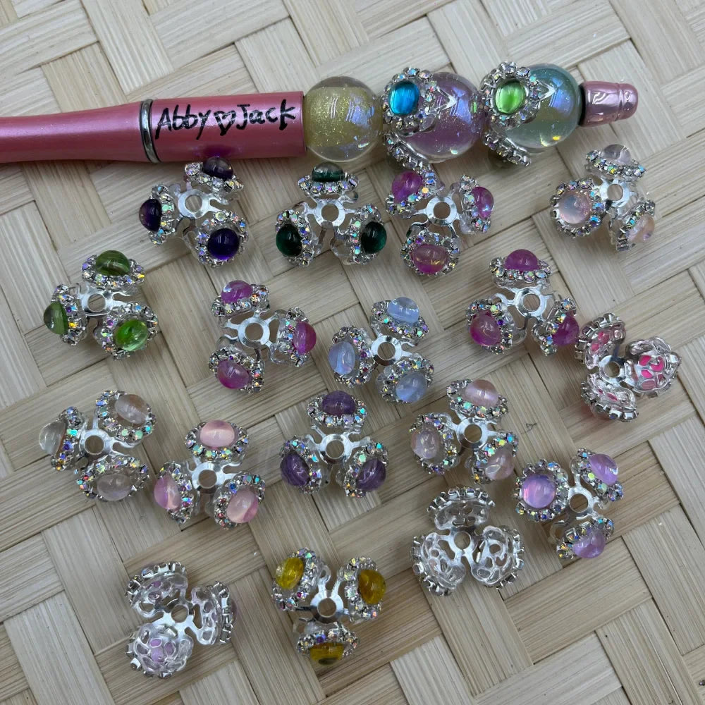 Assorted rhinestone silver bead caps with colorful pastel and jewel-tone cabochon beads in pink, purple, green, yellow, and clear iridescent finishes, displayed on a woven bamboo surface next to a pink beadable pen labeled Abby and Jack.