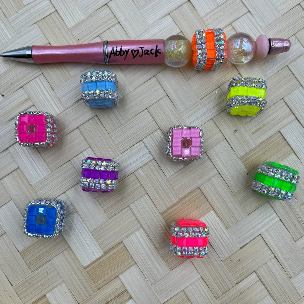 Assorted neon and jewel-toned square fancy beads with rhinestone crystal borders in pink, blue, purple, green, orange, and yellow, displayed next to a beadable pen on a wicker surface.