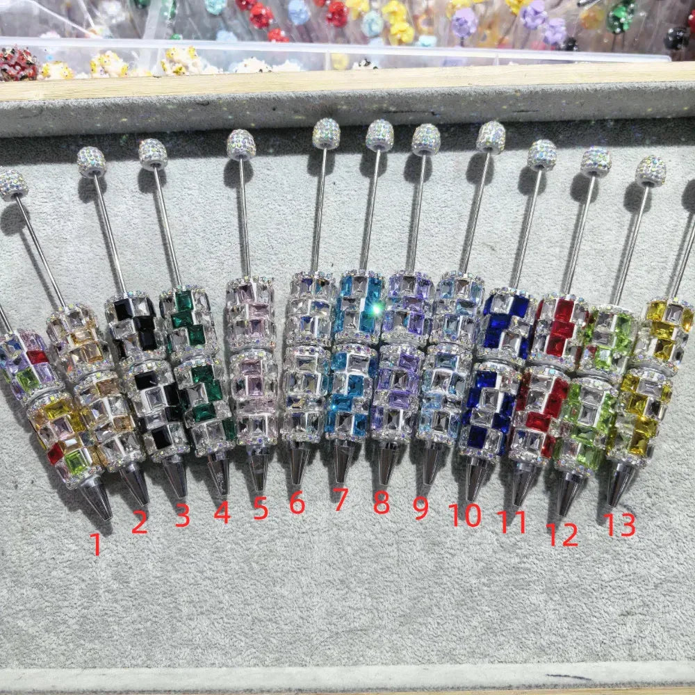 13 crystal rhinestone square gem beadable pens in assorted colors including blue, purple, green, red, yellow, and clear, displayed on gray surface numbered 1 through 13.