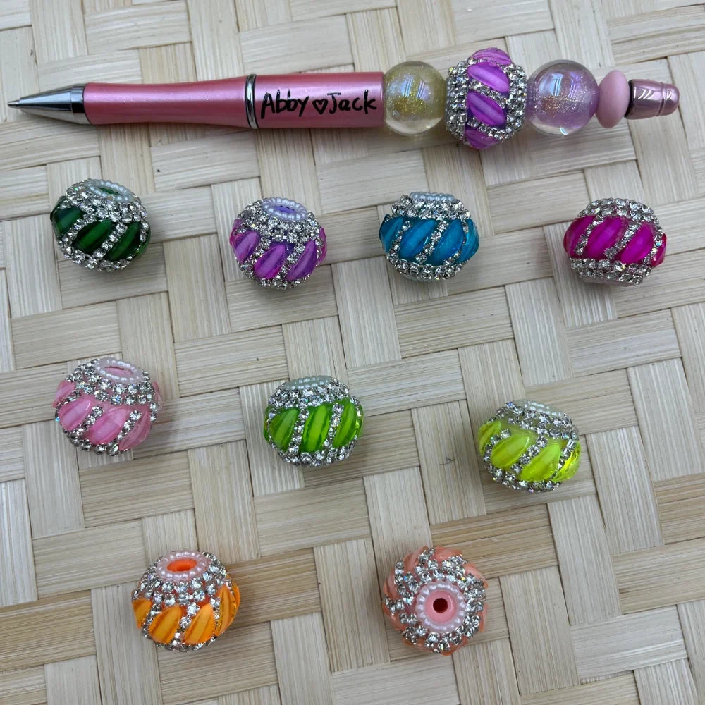 Assorted colorful striped resin beads with rhinestone trim in green, pink, teal, hot pink, orange, and yellow-green, displayed next to a decorative beadable pen on a wicker surface.