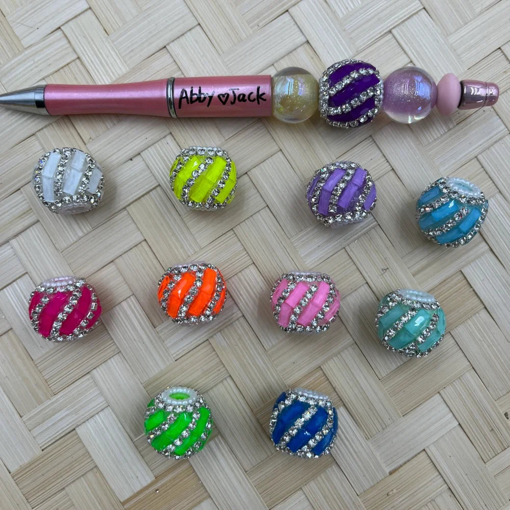 Assorted rhinestone-encrusted striped fancy beads in 10 colors including white, neon yellow, lavender, teal, hot pink, orange, pink, mint, green, and blue, displayed next to a pink beadable pen labeled Abby Jack on a woven bamboo surface.