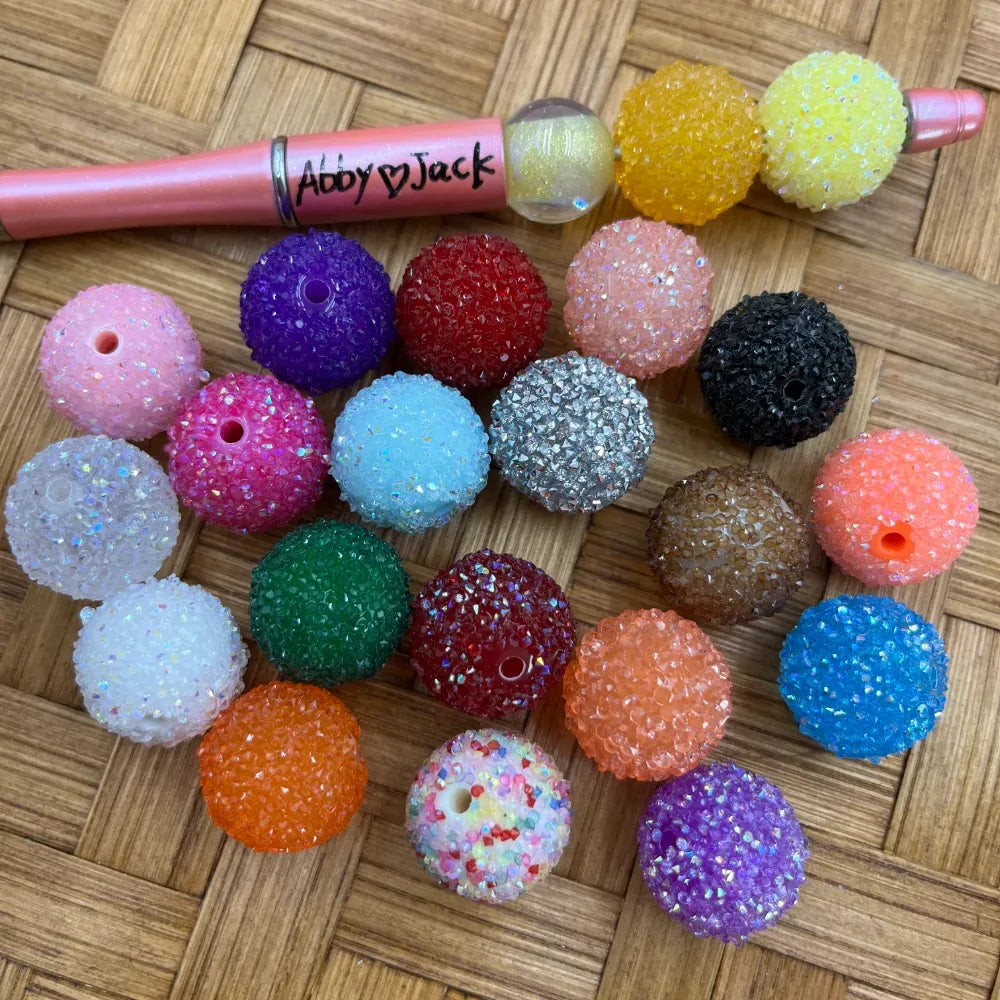 Assorted rhinestone sugar beads in 20+ colors including purple, red, orange, green, silver, black, blue, and multicolor glitter, displayed on a woven surface next to a pink beadable pen labeled Abby and Jack.