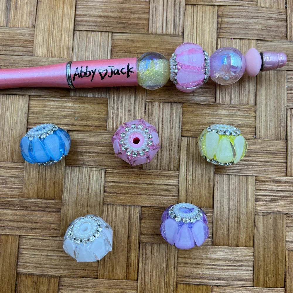 Assorted pastel chunky acrylic fancy beads with rhinestone trim rings in blue, pink, yellow, purple, and white, displayed with a pink beadable pen on a wicker surface.