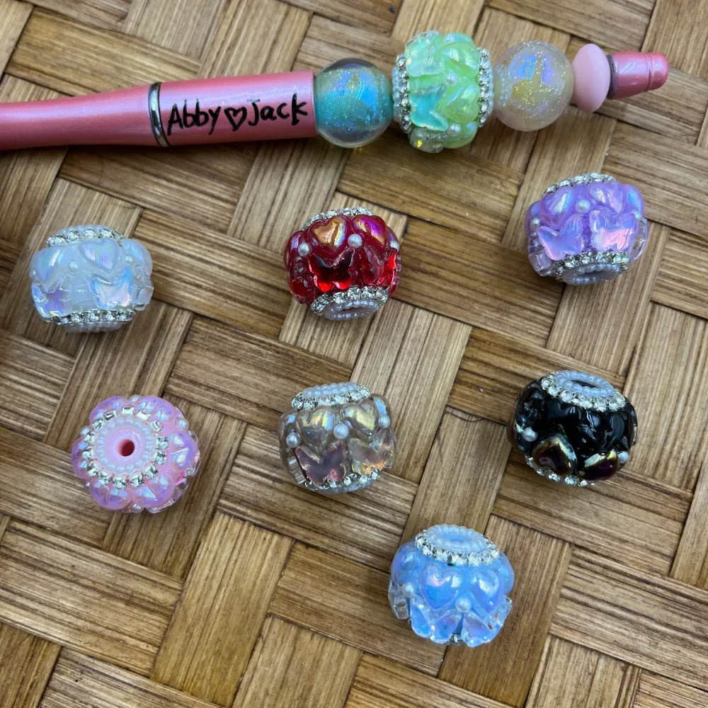 Assorted iridescent rhinestone-trimmed heart floral chunky fancy beads in red, purple, black, blue, white, pink, and clear colors displayed on a wicker surface next to a pink beadable pen labeled Abby and Jack.