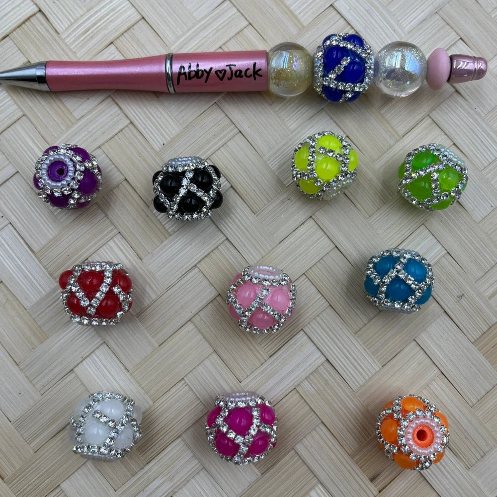 Assorted rhinestone-wrapped chunky round fancy beads in purple, black, yellow, green, red, pink, teal, white, hot pink, and orange displayed on woven surface next to a pink beadable pen labeled Abby Jack.