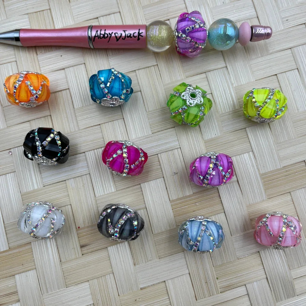 Assorted colorful rhinestone-wrapped melon shaped fancy beads in orange, teal, black, hot pink, purple, green, yellow, white, gray, blue, and pink displayed on woven surface next to a beadable pen.