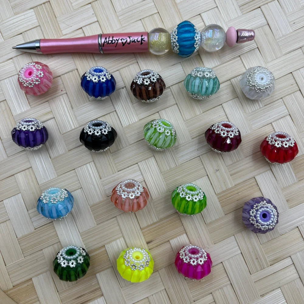 Assorted colorful ribbed melon-shaped glass fancy beads with silver daisy flower caps displayed on a woven surface next to a pink beadable pen, available in pink, blue, brown, teal, purple, black, green, red, yellow, hot pink, and more.