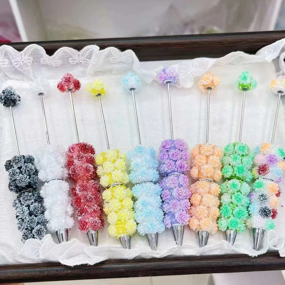 Set of colorful rose flower clay beadable pens arranged in rainbow order including gray, red, yellow, blue, purple, orange and green on a white lace-trimmed tray display.