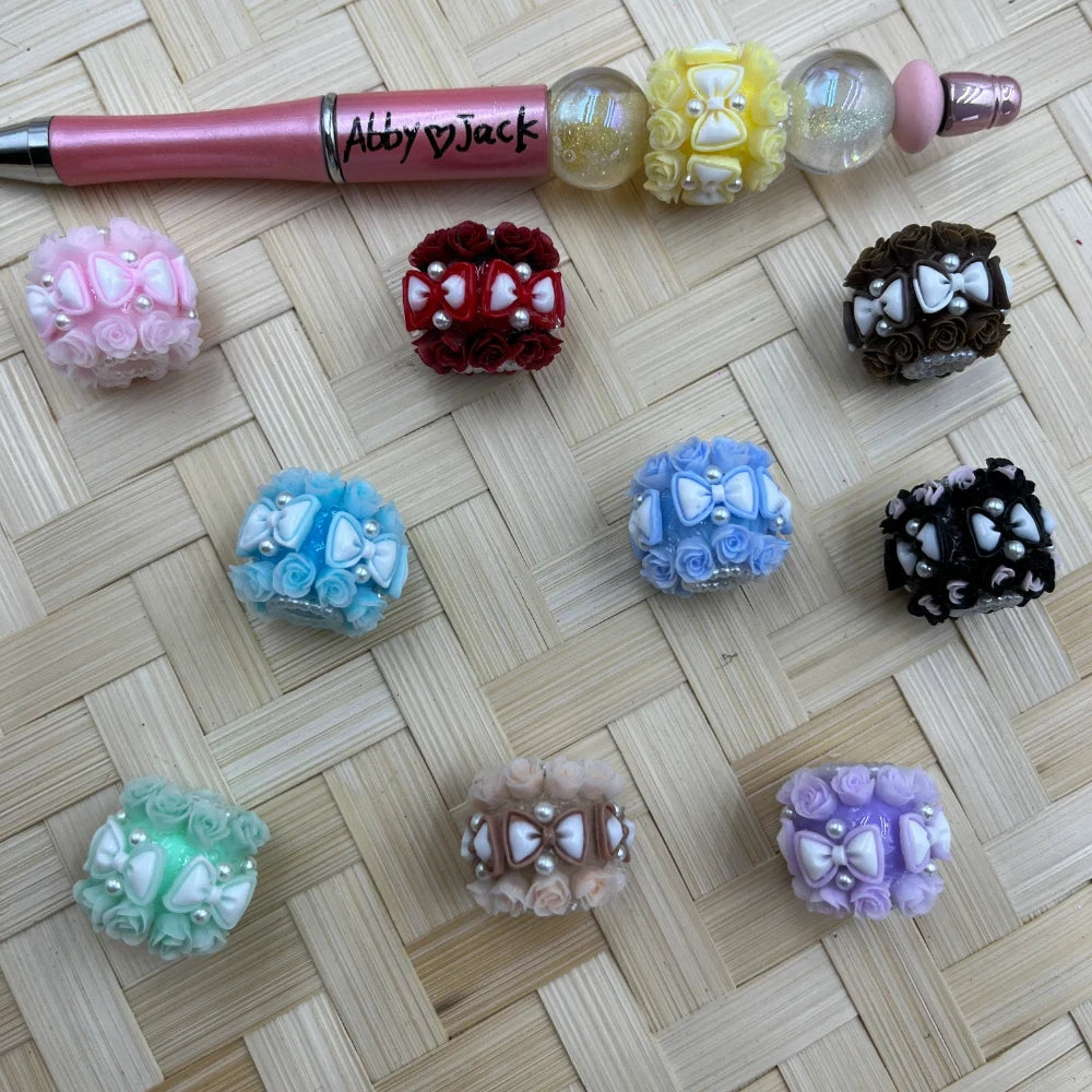Assorted rose floral fancy beads with white bow and pearl accents in 10 colors including pink, red, blue, black, purple, mint, lavender, brown, and yellow, displayed on a woven surface next to a pink beadable pen labeled Abby Jack.
