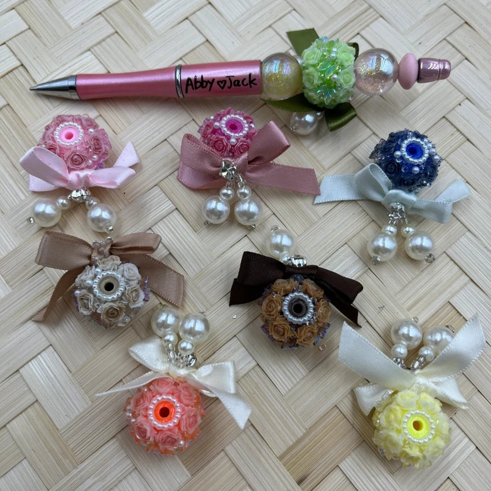 Assorted rose flower ball fancy beads with satin ribbon bows and pearl dangles in pink, navy, brown, coral, and yellow, displayed on woven surface alongside a decorated beadable pen labeled Abby and Jack.