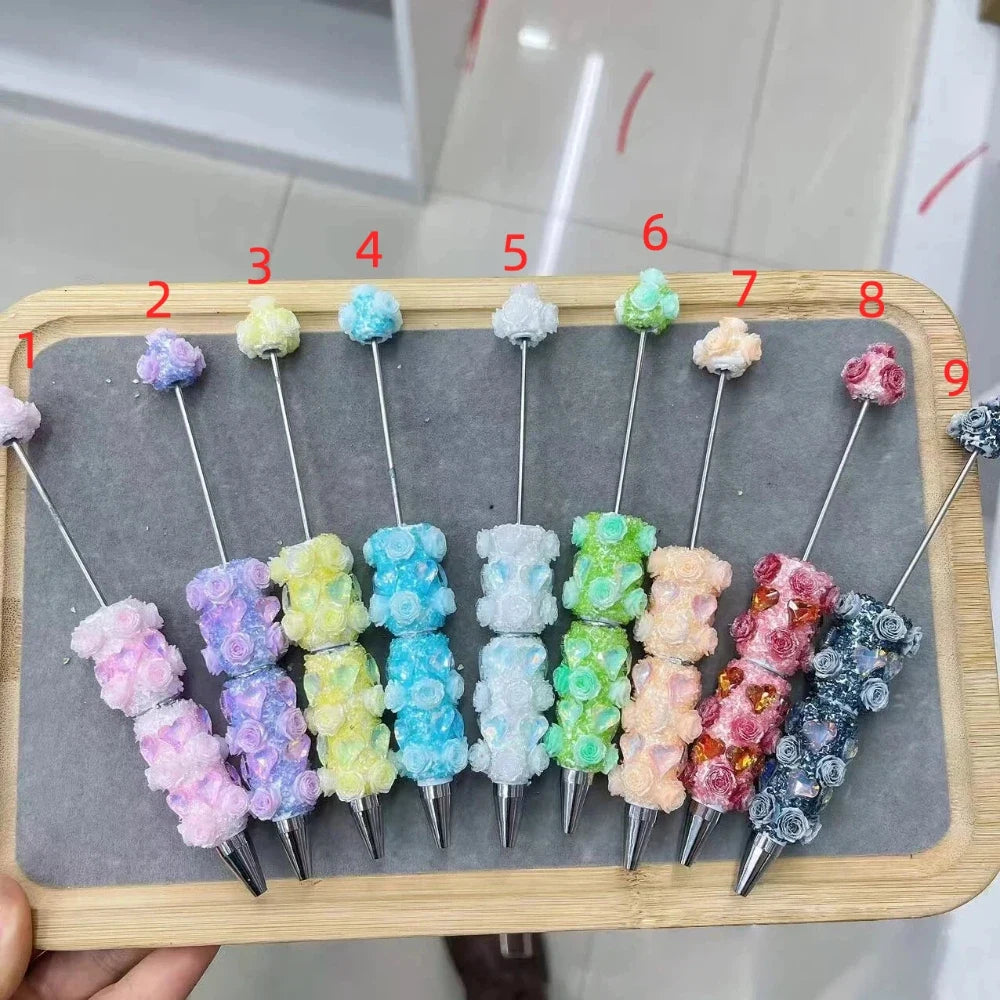 9 colorful rose flower bear clay beadable pens in pastel glitter colors including pink, purple, yellow, teal, white, green, peach, red, and gray displayed on a wooden tray