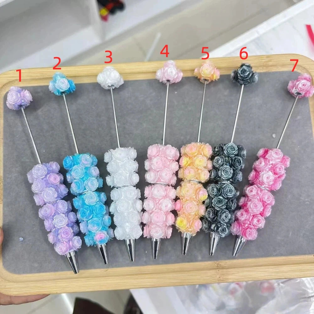 Seven rose flower clay beadable pens displayed on a wooden tray, numbered 1-7, in colors purple, blue, white, pink, peach/yellow, dark gray, and hot pink, each covered in miniature clay roses with a silver metal tip and top bead.