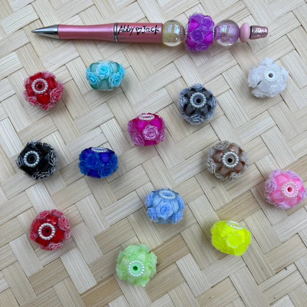 Assorted rose flower cluster resin fancy beads in 14 colors including pink, red, blue, black, green, yellow, and white, displayed on a woven surface next to a beaded pink pen labeled Abby Jack.